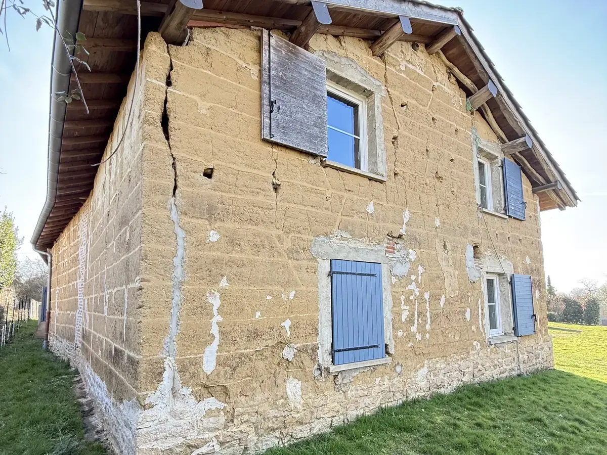 Maison en pisé, enduit tombé par plaques, terre apparente, volets bleus